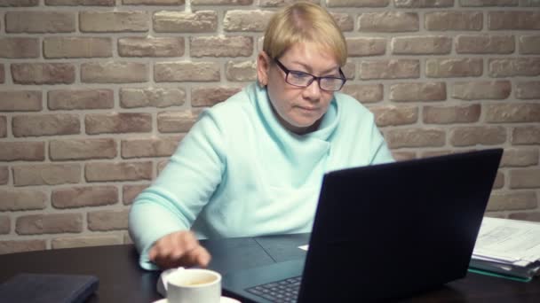 An elderly woman works with a laptop, sitting at a table. elderly work concept — Stock Video