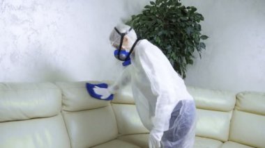 Koruyucu takım elbiseli bir kadın ve solunum cihazı odadaki deri kanepeyi siliyor..