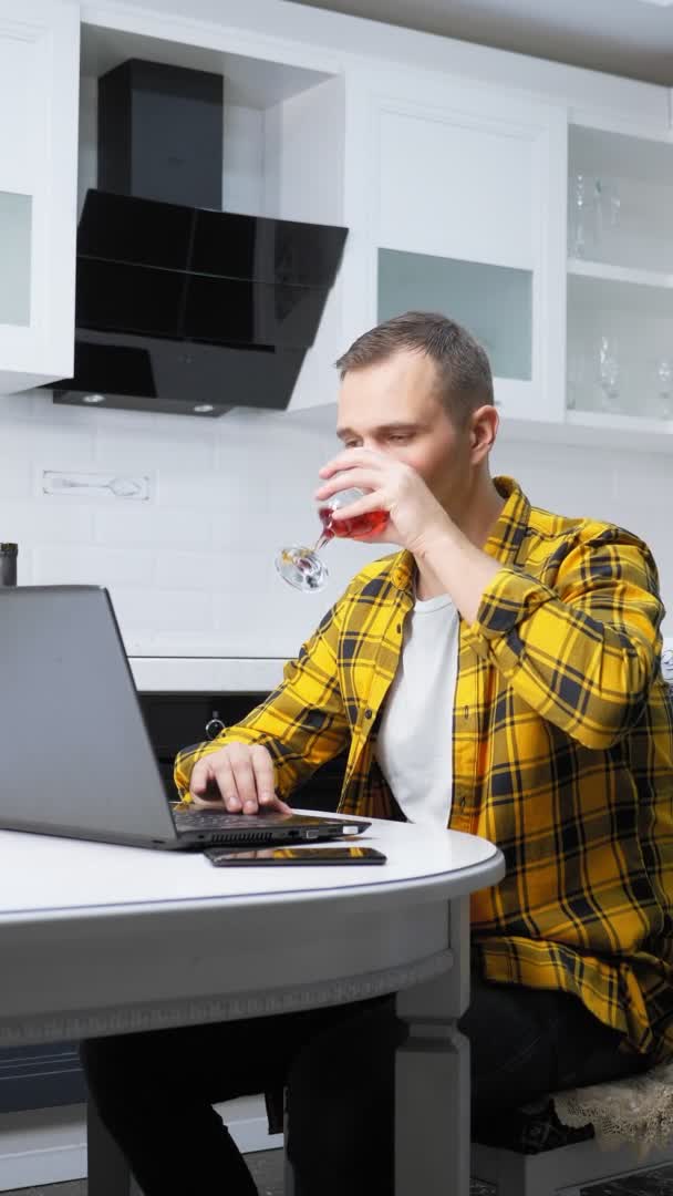 Vertical shot a man in a plaid shirt uses a laptop drinking wine on the kitchen — 图库视频影像