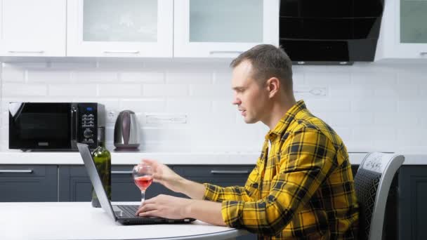 Un hombre con una camisa a cuadros usa un portátil bebiendo vino en la mesa de la cocina — Vídeos de Stock