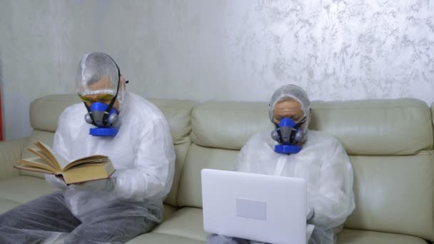 Personas con trajes de protección y respiradores en casa leyendo un libro y usando una computadora portátil — Vídeos de Stock