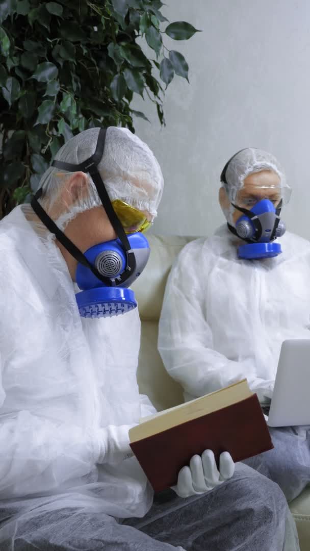 Tiro vertical. pessoas em trajes de proteção e respiradores em casa lendo um livro e usando um laptop — Vídeo de Stock