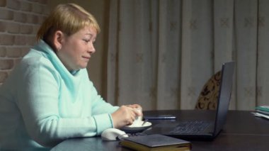 Yaşlı bir kadın dizüstü bilgisayarın önünde otururken videoda konuşuyor.