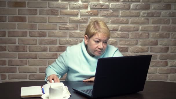 Μια ηλικιωμένη γυναίκα δουλεύει με ένα λάπτοπ, κάθεται σε ένα τραπέζι. έννοια της εργασίας ηλικιωμένων — Αρχείο Βίντεο