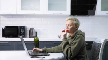 beautiful stylish woman is using laptop and drinking wine ,  in the kitchen