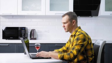 Ekose gömlekli bir adam dizüstü bilgisayar kullanıyor ve mutfak masasında şarap içiyor.