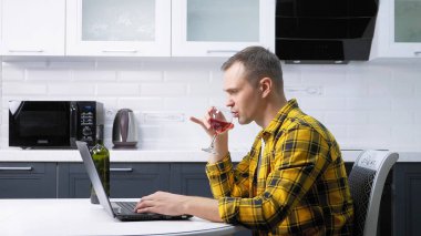 Ekose gömlekli bir adam dizüstü bilgisayar kullanıyor ve mutfak masasında şarap içiyor.