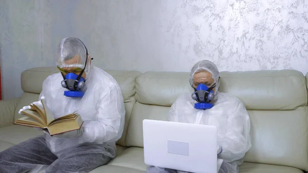 People in protective suits and respirators at home reading a book and using a laptop — Stock Photo, Image