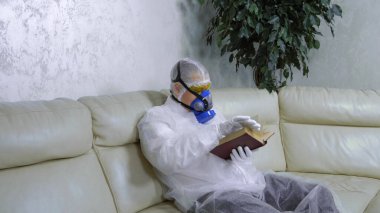 Koruyucu takım elbiseli ve solunum cihazlı bir adam evde kitap okuyor. 