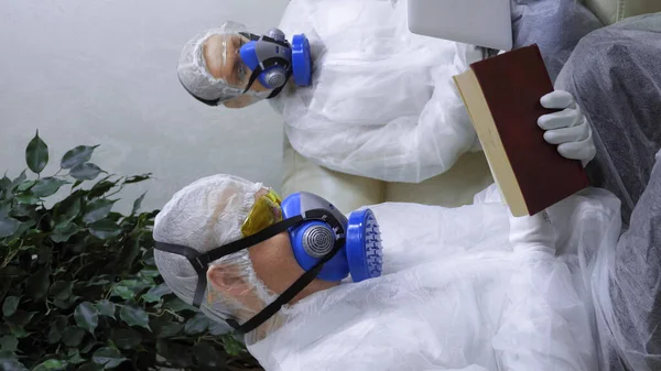 Vertical shot. people in protective suits and respirators at home reading a book and using a laptop — Stock Photo, Image