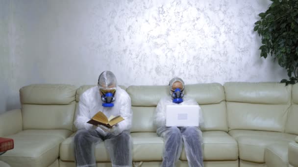 Personas con trajes de protección y respiradores en casa leyendo un libro y usando una computadora portátil — Vídeos de Stock