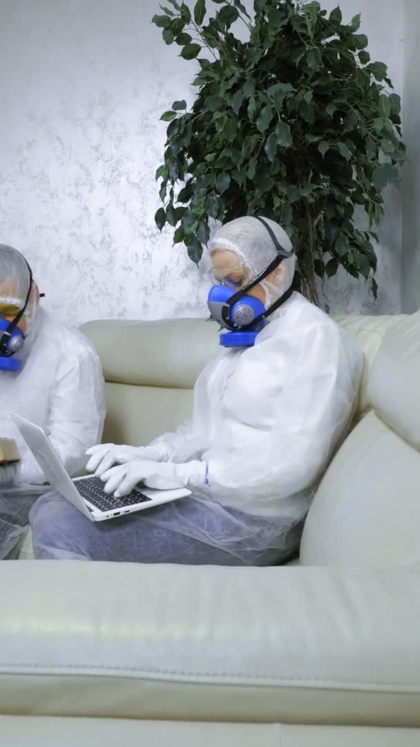 Personas con trajes de protección y respiradores en casa leyendo un libro y usando una computadora portátil. tiro vertical — Vídeo de stock