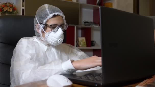 Solunum cihazlı ve koruyucu giysili adam evde otururken laptopunu kullanıyor. — Stok video