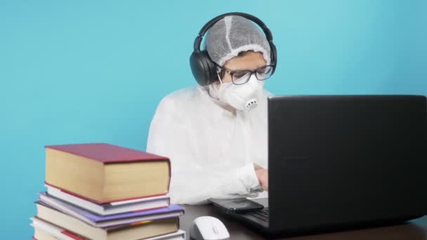 Een man met een beschermend masker en pak gebruikt een laptop en boeken. blauwe achtergrond — Stockvideo