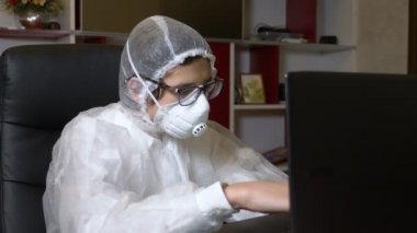 Solunum cihazlı ve koruyucu giysili adam evde otururken laptopunu kullanıyor.
