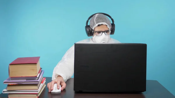 A guy in a protective mask and suit uses a laptop and books. blue background — Stock Photo, Image