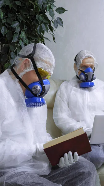 People in protective suits at home reading a book, using a laptop. vertical shot — Stock Photo, Image