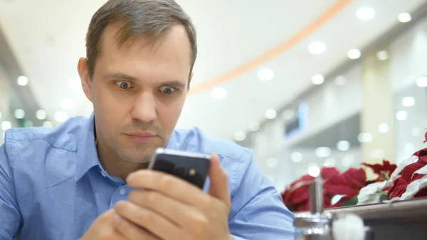 Joven hombre guapo sorprendido cuando se utiliza un teléfono inteligente mientras está sentado en un café — Foto de Stock