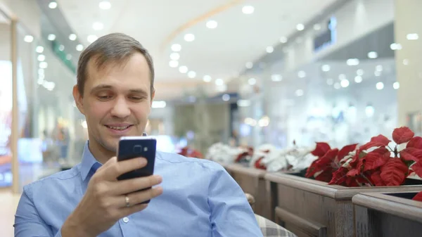 Joven guapo está utilizando el teléfono inteligente sentado en la cafetería, sonriendo . — Foto de Stock