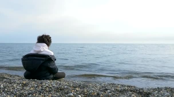 Adolescente niño sentado solo en la playa en tiempo frío y mirando hacia el mar — Vídeo de stock