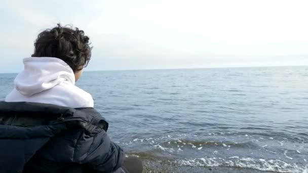 Adolescente niño sentado solo en la playa en tiempo frío y mirando hacia el mar — Vídeo de stock