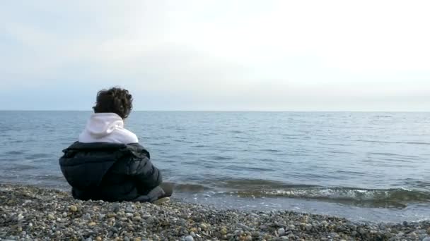 Хлопчик сидить один на пляжі в холодну погоду і дивиться в море — стокове відео