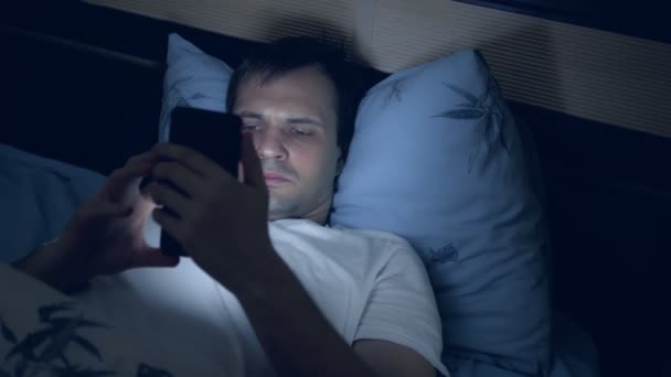 Un joven usa un teléfono inteligente en la cama por la noche. insomnio — Vídeo de stock