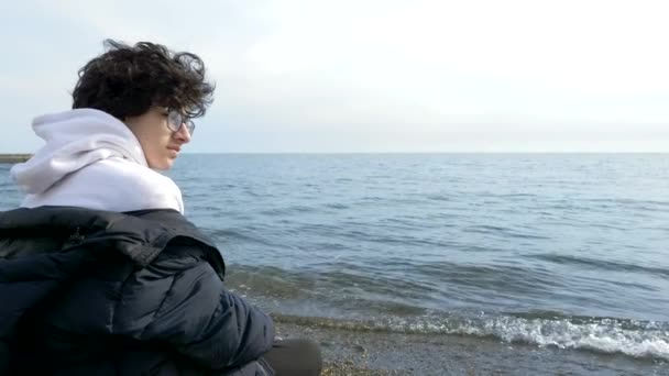 Genç çocuk soğuk havada sahilde tek başına oturuyor ve denize bakıyor. — Stok video