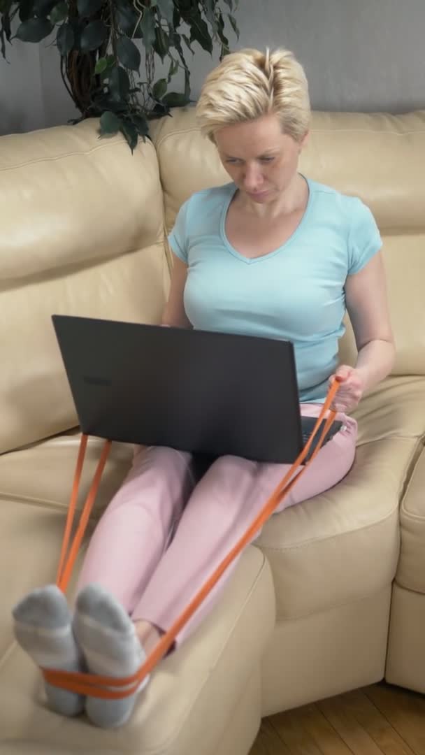 Η γυναίκα ασχολείται με τον αθλητισμό στο σπίτι με ένα ελαστικό ιμάντα και χρησιμοποιεί το φορητό υπολογιστή — Αρχείο Βίντεο