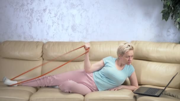 La femme fait du sport à la maison avec une bande élastique et utilise l'ordinateur portable — Video