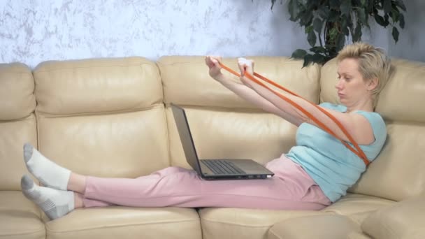 Женщина занимается спортом дома с эластичной лентой и использует ноутбук — стоковое видео