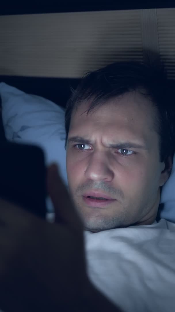 Κάθετη λήψη ενός νεαρού άνδρα χρησιμοποιεί ένα smartphone στο κρεβάτι τη νύχτα — Αρχείο Βίντεο