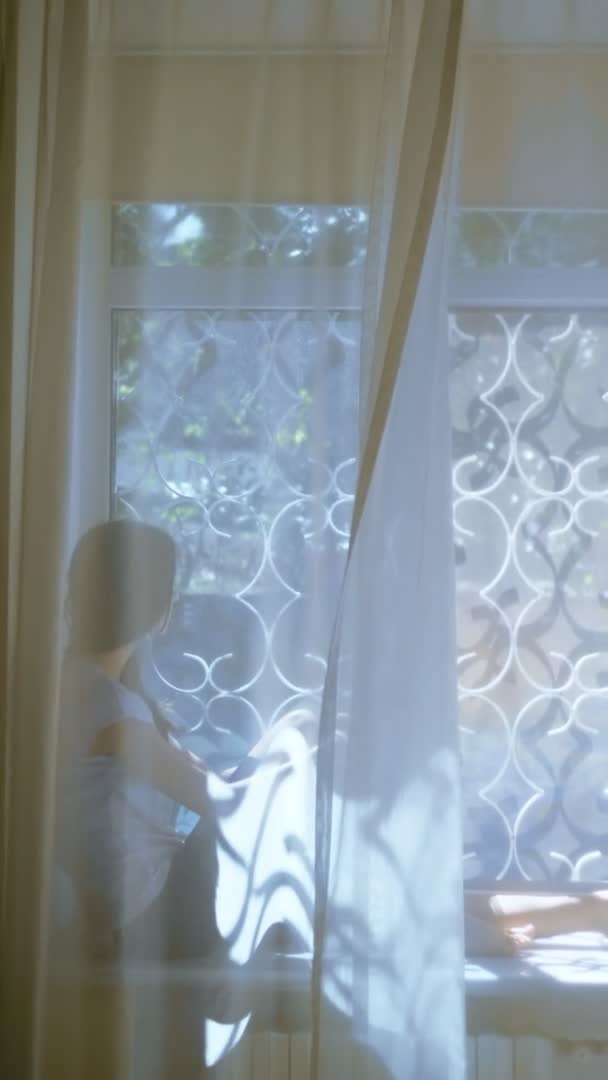 Вертикально. дівчина-підліток сидить на підвіконні і дивиться у вікно. розмивання — стокове відео