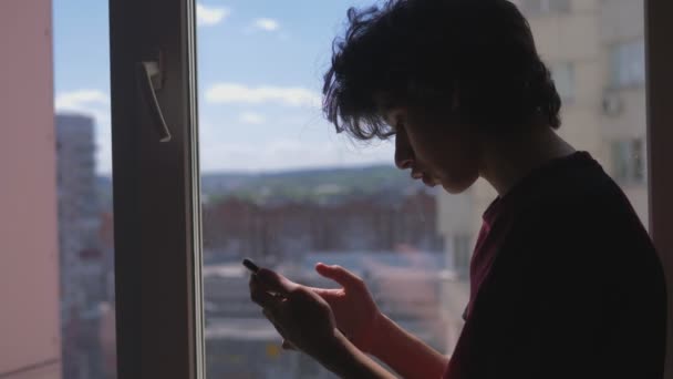 Silueta. Apropiere. adolescent băiat folosește un smartphone stând pe pervazul ferestrei — Videoclip de stoc