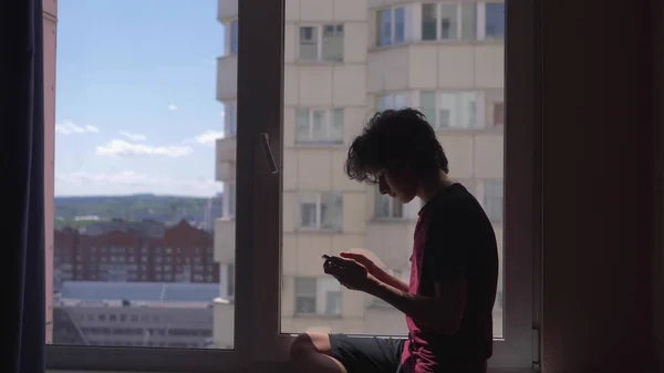 Silhueta. adolescente usa smartphone sentado no peitoril da janela em seu quarto — Fotografia de Stock