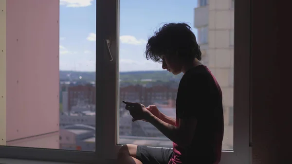 Silueta. adolescente utiliza teléfono inteligente sentado en alféizar ventana en su habitación —  Fotos de Stock