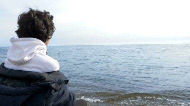 Genç çocuk soğuk havada sahilde tek başına oturuyor ve denize bakıyor.
