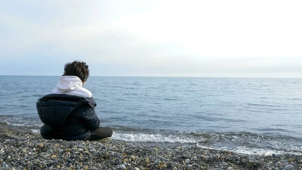Genç çocuk soğuk havada sahilde tek başına oturuyor ve denize bakıyor.