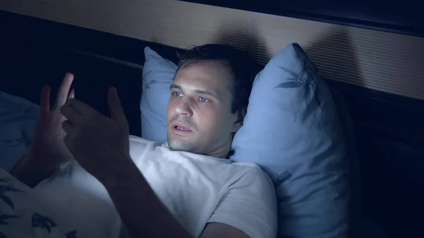 Młody mężczyzna w nocy używa smartfona w łóżku. bezsenność — Zdjęcie stockowe