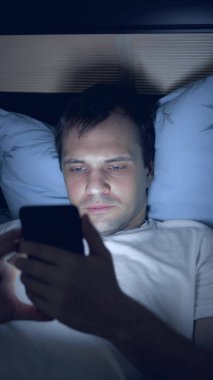 Yatakta akıllı telefon kullanan genç bir adamın dikey görüntüsü.