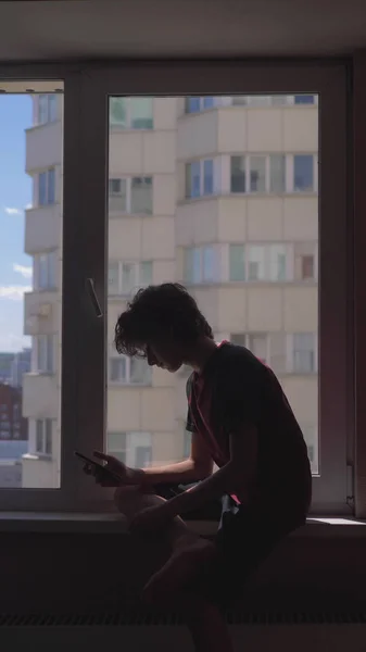 vertically. silhouette. teenage boy uses a smartphone sitting on the window sill