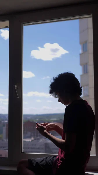 Verticalmente. silhueta. adolescente usa um smartphone sentado no peitoril da janela — Fotografia de Stock