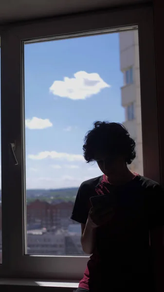 Pionowo. sylwetka. nastolatek chłopiec używa a smartfon siedzi na w okno sill — Zdjęcie stockowe