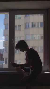 Dikey olarak. Silüet. Genç çocuk pencere eşiğinde akıllı telefon kullanıyor.