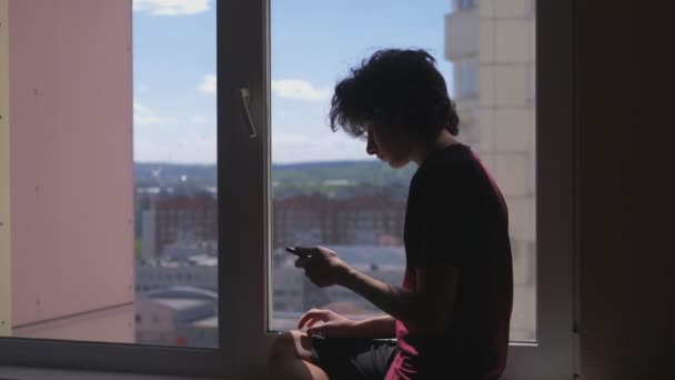 Silhouette. teenage boy uses smartphone sitting on window sill in his room — Stock Video