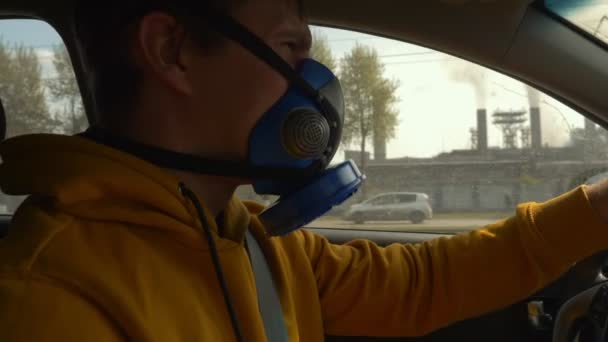 Een man met een masker rijdt in een auto langs de rokende schoorstenen van een fabriek. — Stockvideo