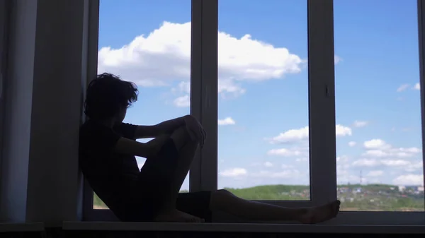 Silhueta. adolescente senta-se em um peitoril da janela contra um céu azul e nuvens brancas — Fotografia de Stock