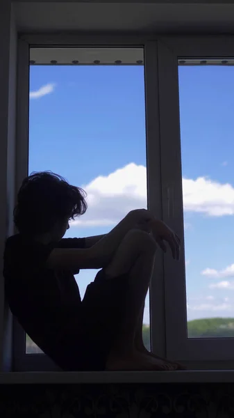 Verticalmente. silhueta. adolescente senta-se em uma janela contra um céu azul — Fotografia de Stock