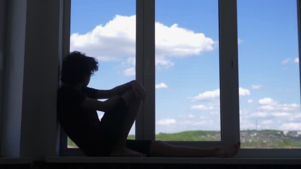 Silhouette. Teenager sitzt auf Fensterbank vor blauem Himmel und weißen Wolken — Stockvideo