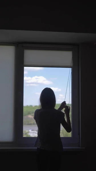 vertically. silhouette. girl opens the blinds, view of the sky